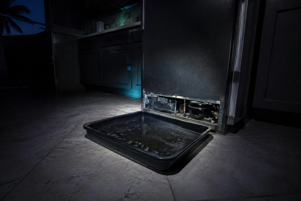 A shallow black drain pan slid out from underneath the base of a refrigerator that stands pulled forward on a tile floor, a little dirty water still lying in it.