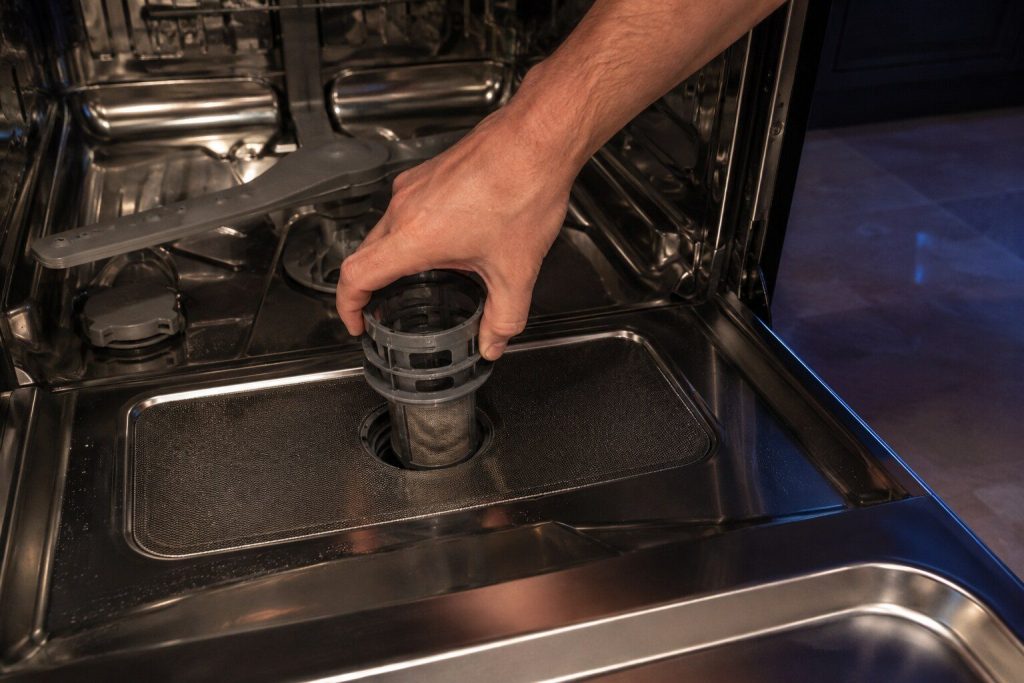A hand turning the cylindrical filter counter-clockwise out of the floor of a dishwasher tub, the flat mesh plate still in place beneath it.