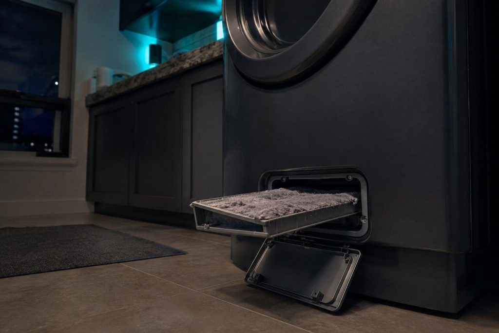 A rectangular lint filter drawn out of an open hatch at the bottom front of a ventless dryer, packed grey with lint, the hatch door hanging down.