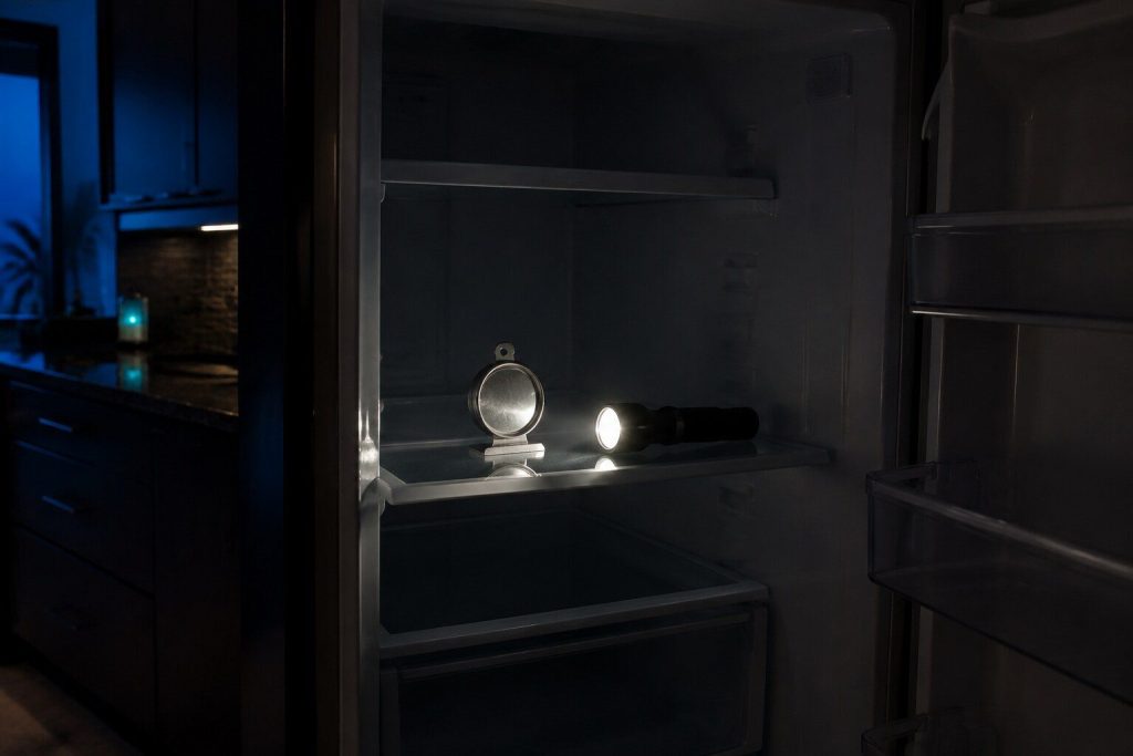 An appliance thermometer standing on a refrigerator shelf photographed by torchlight with the interior lamp off and the compartment dark, a torch lying on the shelf beside it.