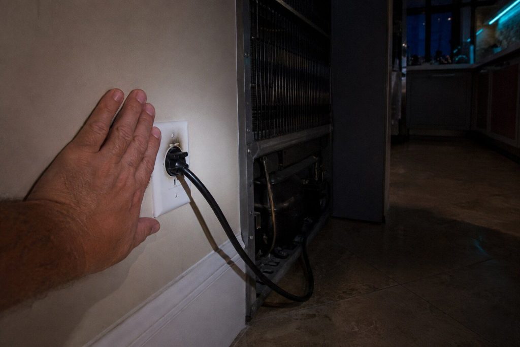 The back of a hand held against the faceplate of a wall outlet behind an appliance pulled out from the wall, the plug seated in the socket beside it, a scorch shadow on the plate.