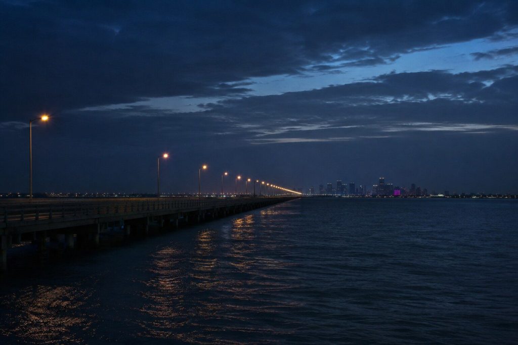 A causeway across dark water at dusk with a line of lamps running away into the distance and a city skyline low on the horizon.