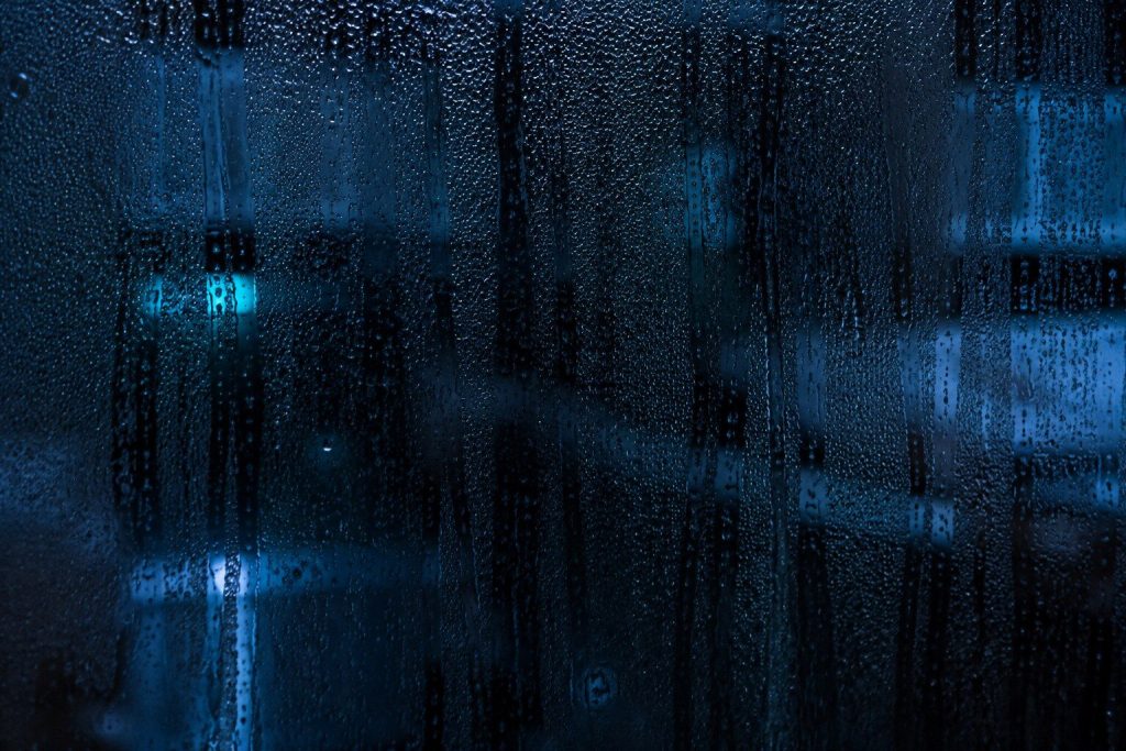 Condensation running down the inside of a dark kitchen window at night, the room behind it out of focus and blue.
