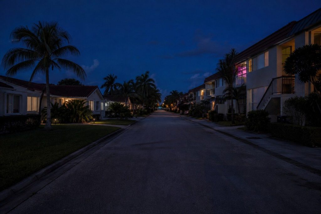 A South Florida residential street at blue hour seen down its length, low buildings and palms either side, warm windows just coming on and the sky deep blue above.