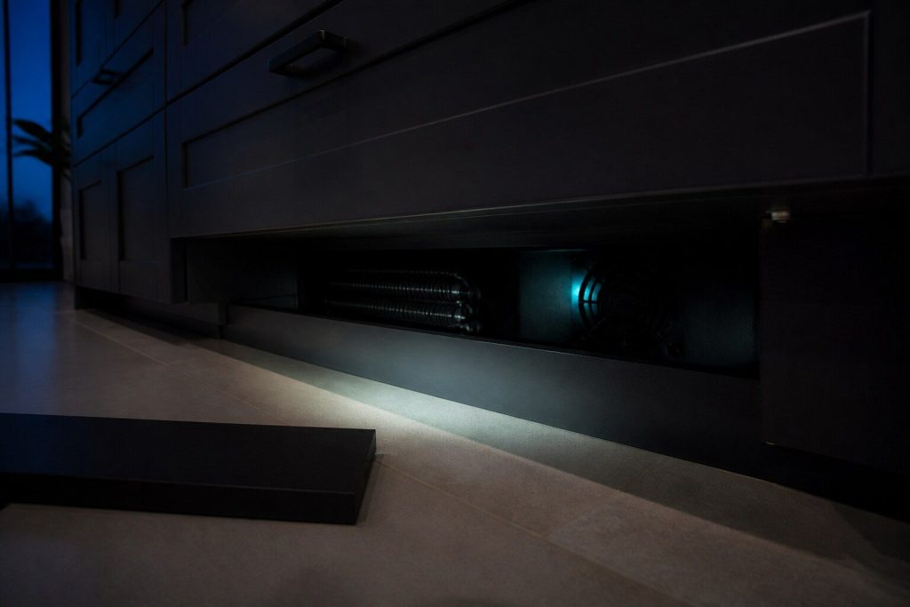 The dark recess behind a removed kickboard at the base of a run of kitchen cabinets, a coil and a fan grille just visible in the shadow.