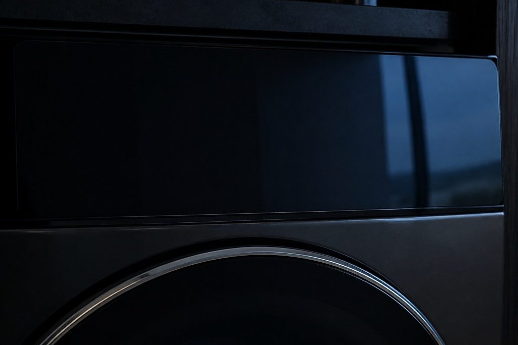 The black glass face of an appliance control panel filling the frame, unlit, carrying only a soft reflection of a window.