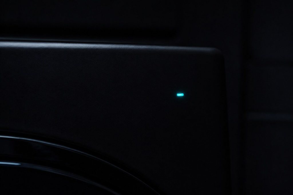 One small indicator lamp glowing on an otherwise completely dark appliance panel in an unlit room, everything around it black.