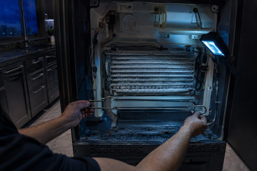 A tubular defrost heater lifted clear of the bottom of an evaporator coil and held horizontal, the finned coil behind it still carrying frost, the back panel off.