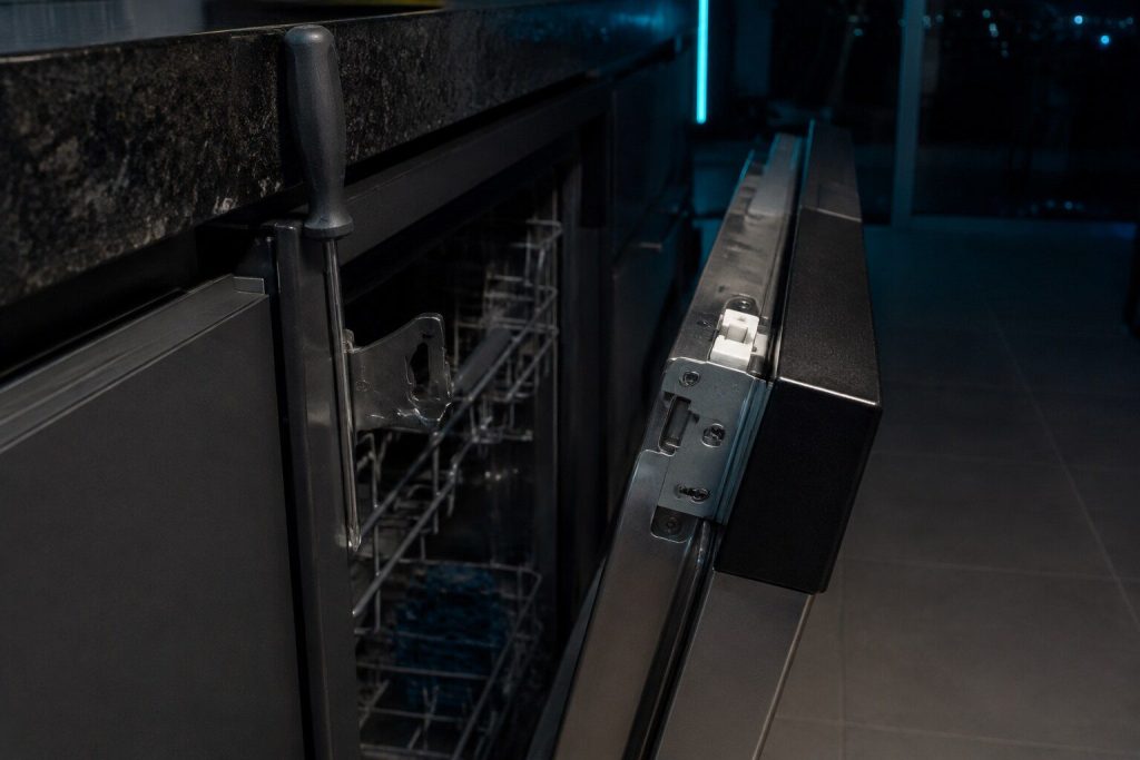 The latch assembly at the top of a dishwasher door and the strike it closes into, the door held an inch open so both halves are visible, a small screwdriver resting against the frame.