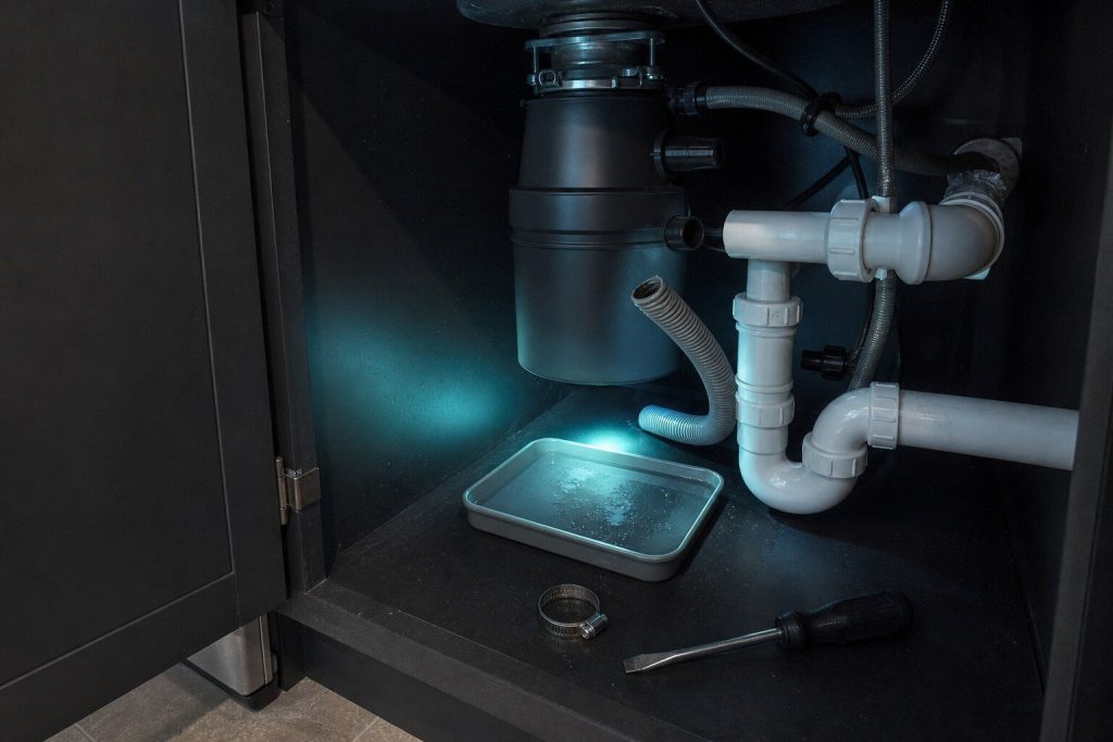 A corrugated drain hose pulled off a waste disposer spigot under a sink with a shallow tray beneath catching the water, a hose clamp and a screwdriver lying on the cabinet floor.