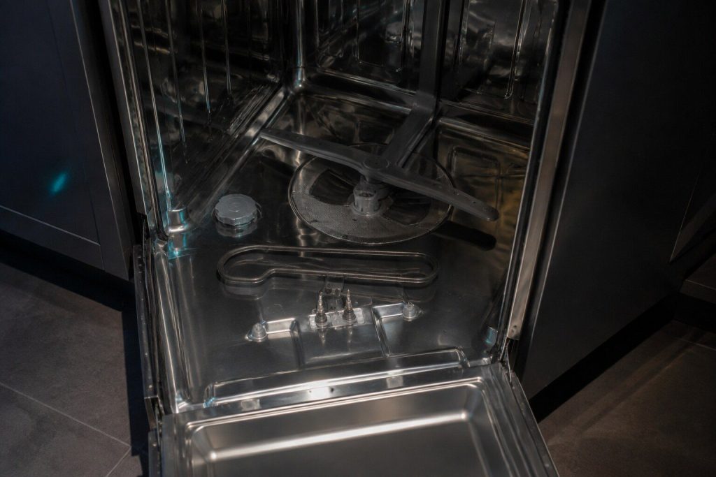 The looped heating element lifted a little clear of the floor of a dishwasher tub with the lower rack out, its two terminals visible where they pass through the base.