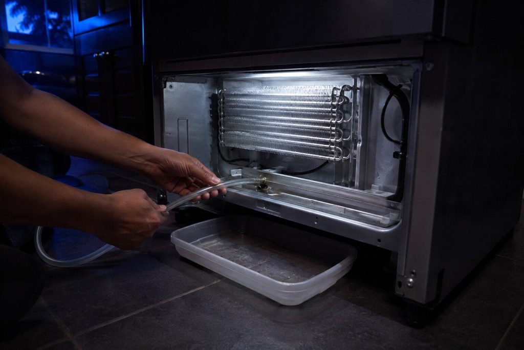 A length of flexible tubing being fed into the drain trough at the bottom of an evaporator compartment, the back panel off and a shallow tray under the machine to catch what comes through.