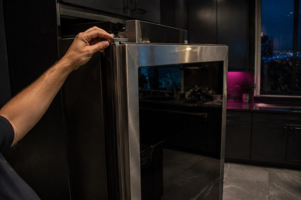 A refrigerator door with a large dark glass panel set into its front, held half open while a hand checks the hinge at the top of the door.