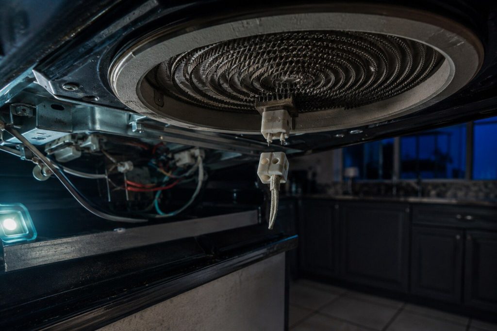 A ceramic connector unplugged from the tail of a radiant cooktop element seen from underneath the lifted glass top, the coil of the element visible above it.