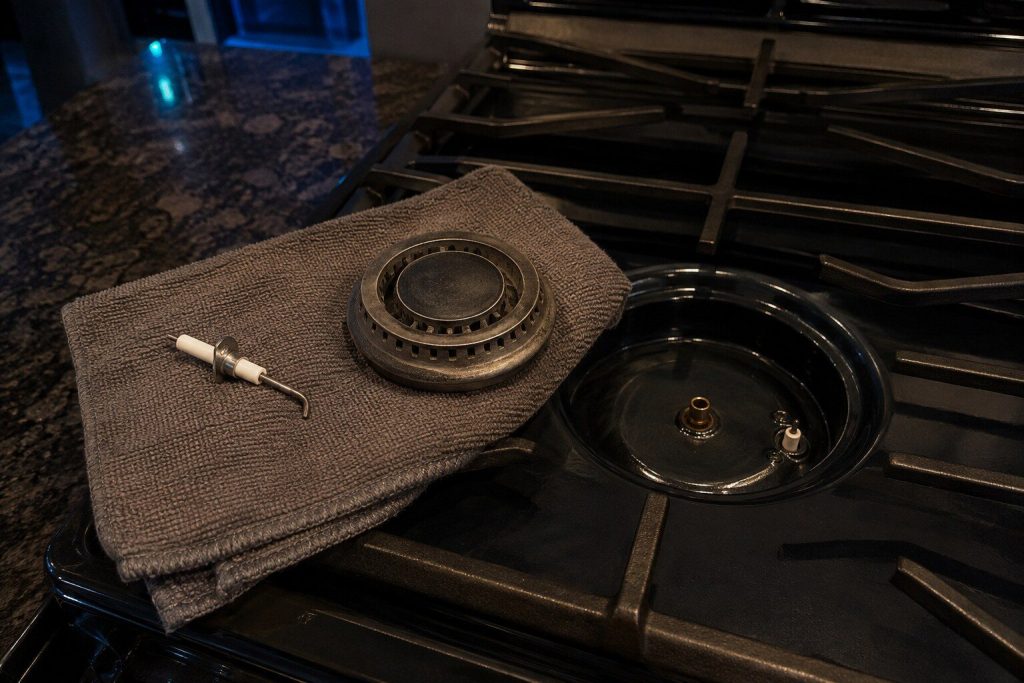A spark igniter and a burner head lying apart on a folded towel on a cooktop, the empty burner base and its orifice visible beside them.