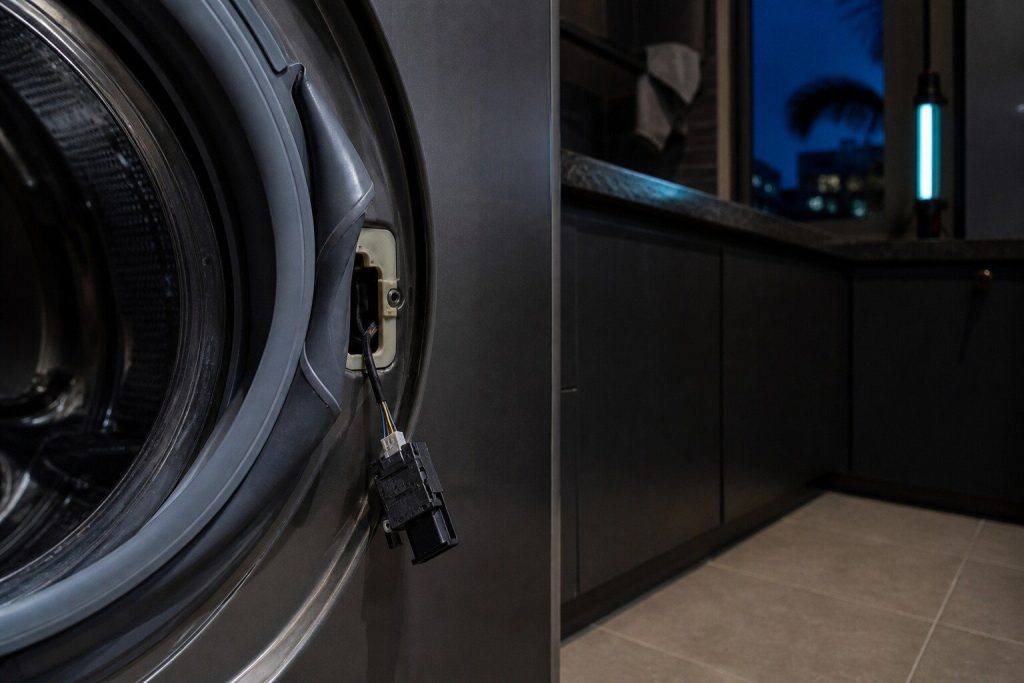 A door lock unit drawn out of its cutout in the front panel of a washing machine and hanging on its wiring loom, the gasket folded aside.