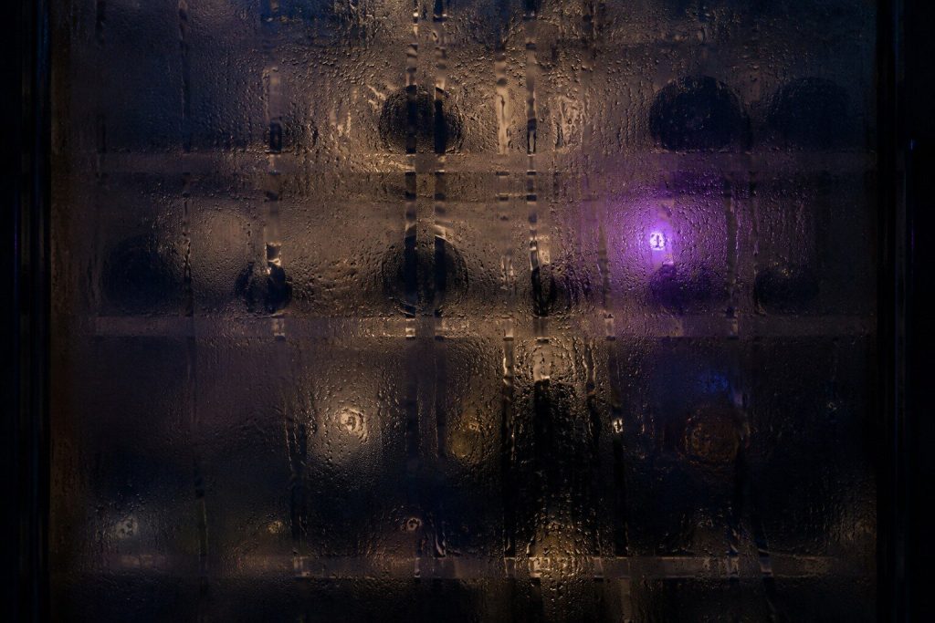 A sheet of heavy condensation on the INSIDE of a wine cabinet's glass door, thick enough that the bottles behind it are only dark blurred shapes and beads of water are running down the glass in tracks.