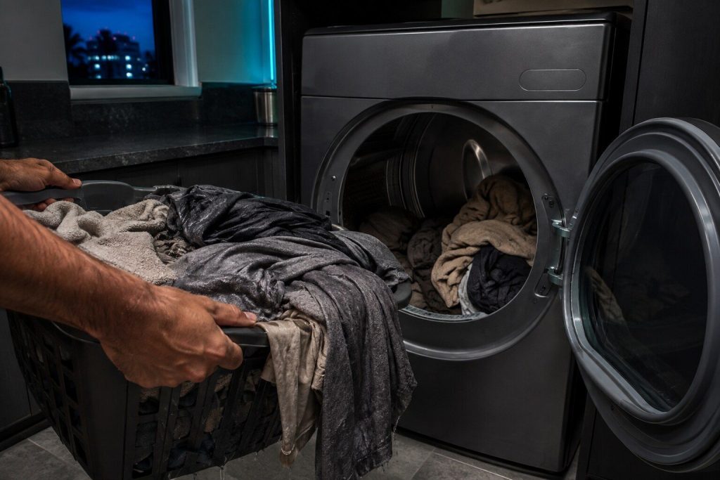 A basket of clothes lifted out of a dryer that still hang heavy and damp, the drum behind still half loaded, the door open.
