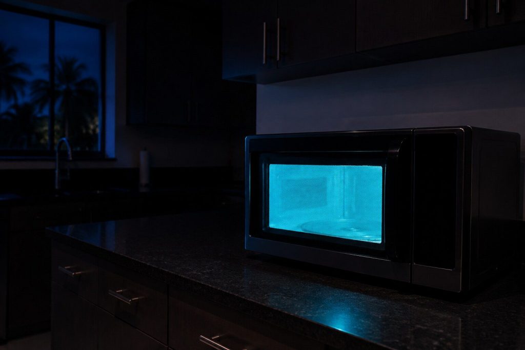 A countertop microwave running by itself in an otherwise dark kitchen at night, its cavity lit and its turntable turning, nobody in the room, the rest of the counter in shadow.