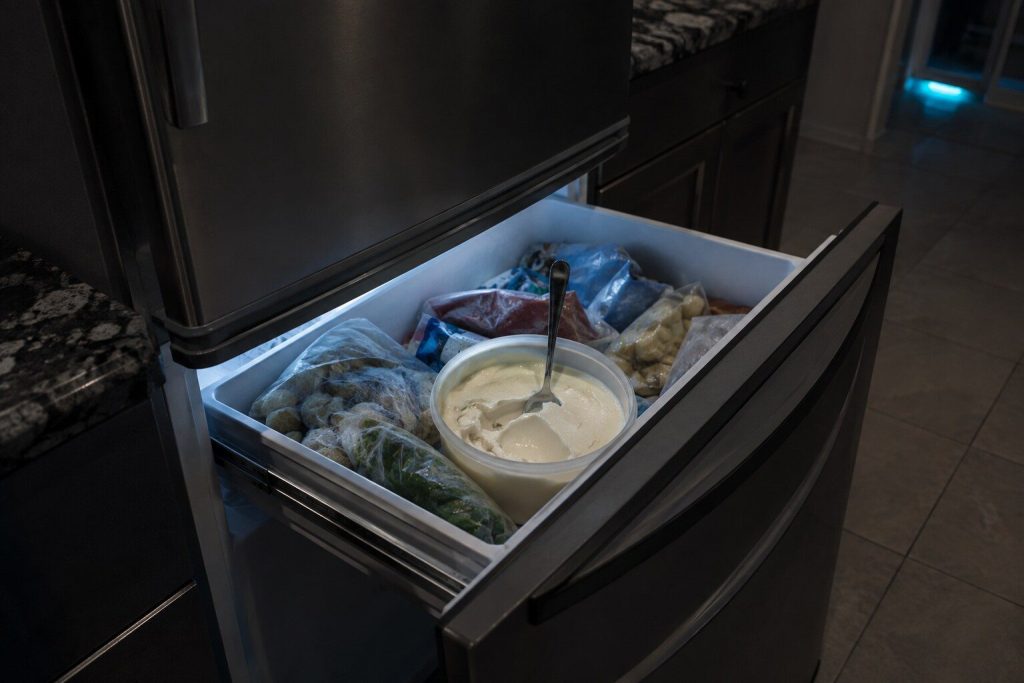 An open tub of ice cream gone soft and glossy in a freezer drawer, a spoon standing upright in it, the packets around it slumped rather than rigid.