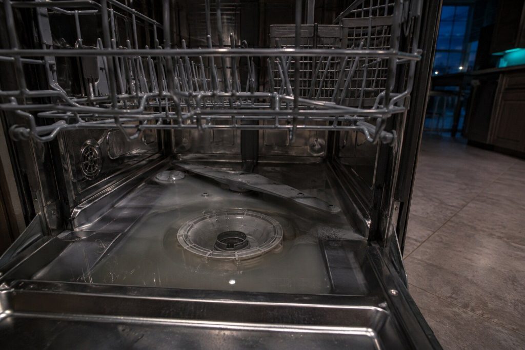 Grey standing water lying in the bottom of an open dishwasher tub around the filter housing, the lower rack pulled half out above it.
